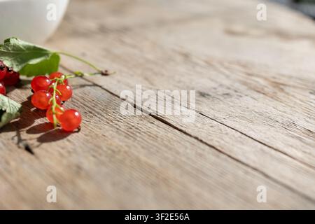 Grappolo di ribes rossi lucidi con foglie verdi poggiato su tavolo di legno consumato, bacche a fuoco netto, luce soffusa del giorno, sfondo sfocato, texture naturale, atmosfera di raccolta fresca, quiete calda di campagna Foto Stock