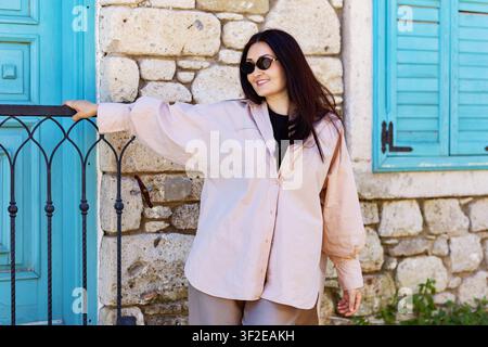 Donna elegante con occhiali da sole si appoggia a una recinzione in ferro battuto accanto a un muro di pietra rustico con porta e persiane blu brillante, godendosi una giornata soleggiata in una cha Foto Stock