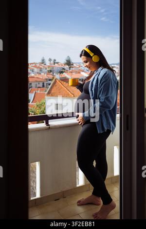 Donna incinta con cuffie gialle si gode un caffè mattutino su un balcone soleggiato, tiene la pancia mentre guarda i tetti rossi e una città tranquilla Foto Stock