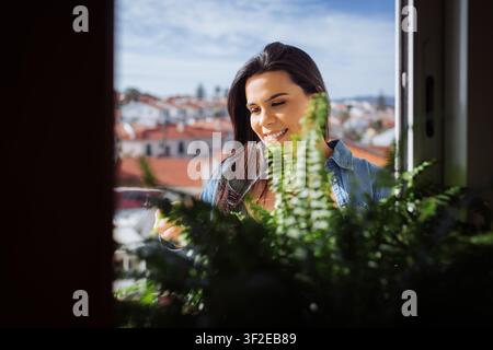Donna felice in una camicia di jeans casual che annaffia felci verdi rigogliose su un balcone di un appartamento, godendosi il sole con una skyline cittadina e tetti sfocati Foto Stock