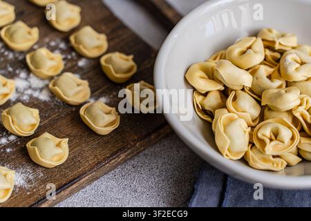 Primo piano di tortellini fatti a mano su una tavola di legno infarinata accanto a una ciotola bianca ripiena di gnocchi freschi, stilizzati come cucina italiana rustica e Foto Stock