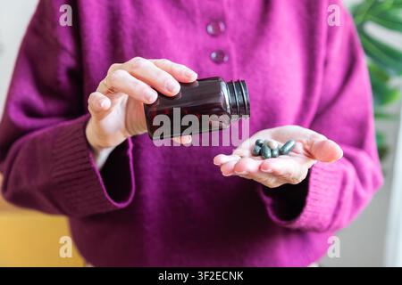 Primo piano di una donna anziana a casa che versa capsule di integratori nella sua mano, sostenendo una routine quotidiana di benessere, invecchiamento sano, cura di sé e preve Foto Stock