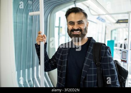 Uomo indiano sorridente in piedi in un treno moderno, che tiene un palo con una mano e indossa un blazer e uno zaino, rappresentando il pendolarismo urbano, i viaggi, Foto Stock