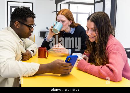 Tre colleghi si radunano intorno a un tavolo giallo in un ufficio moderno, sorridendo mentre guardano uno smartphone e sorseggiano caffè da tazze durante un tè rilassato Foto Stock