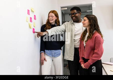Tre giovani colleghi collaborano in un ufficio moderno, rivedono post-it colorati su una parete durante una sessione di pianificazione agile, sorridendo e discutendo Foto Stock