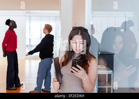 Studenti multietnici si riuniscono in un corridoio luminoso del campus durante una pausa caffè, uno controlla uno smartphone mentre tiene un quaderno e i compagni di classe chiacchierano Foto Stock