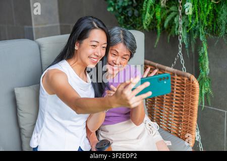Madre asiatica e figlia adulta sorridono mentre scattano un selfie con lo smartphone insieme, mostrando il legame familiare, l'uso della tecnologia e la cura degli anziani. Foto Stock