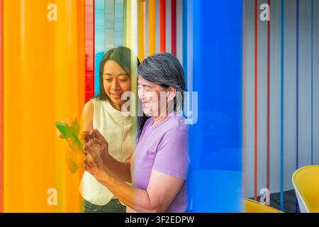 Madre asiatica anziana e figlia adulta sorridono mentre usano insieme un tablet digitale, imparando la tecnologia all'interno con pannelli traslucidi colorati, fami Foto Stock