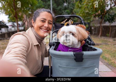 Una donna scatta un selfie sorridendo con i suoi cani, comodamente seduta in un passeggino durante una passeggiata tranquilla in un parco a Taiwan, circondata da una vegetazione lussureggiante G Foto Stock