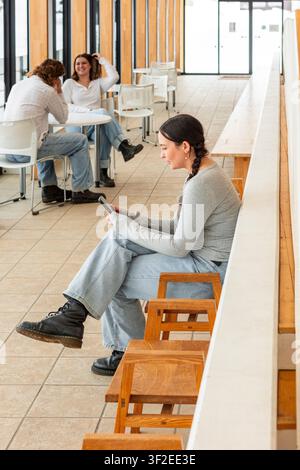 Gli studenti fanno una pausa in una luminosa e moderna lounge del campus. Una è seduta e usa il telefono mentre altri due chiacchierano in sottofondo, godendosi il loro tempo libero Foto Stock