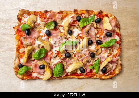 Pinsa romana italiana quattro stagioni ricoperta di pomodoro, mozzarella, prosciutto, funghi, carciofi, olive nere e basilico fresco, presentata su legno Foto Stock