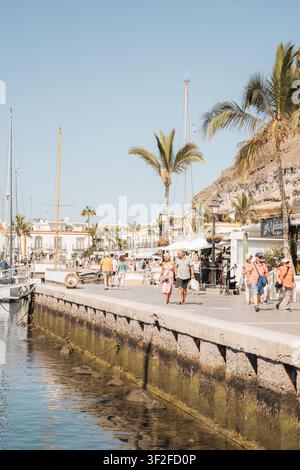 Puerto de Mogan, Spagna – 17 luglio 2025: Vista della passeggiata del porto con palme, persone che camminano, barche ormeggiate nell'acqua e scogliere rocciose nel Foto Stock
