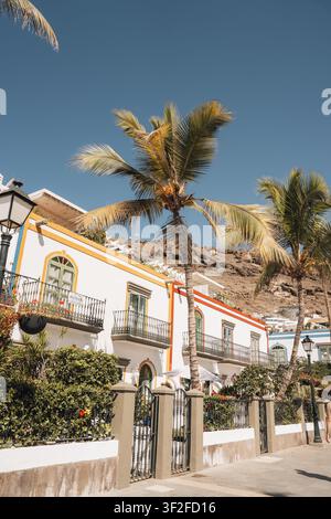 Las Palmas, Spagna - 17 luglio 2025: Vista di edifici bianchi con rifiniture gialle e palme a Puerto de Mogan, con balconi e colline rocciose sotto Foto Stock