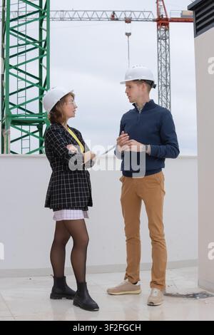 Due giovani professionisti. Un uomo e una donna. Entrambi indossano caschi bianchi. Impegnati in una discussione seria su un tetto che si affaccia su una costruzione attiva Foto Stock