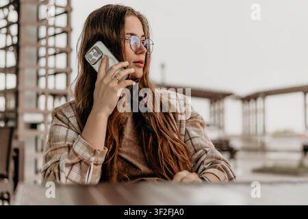Donna, comunicazione smartphone, giovane con occhiali e camicia a quadri che parla al telefono cellulare in un caffè di strada Foto Stock