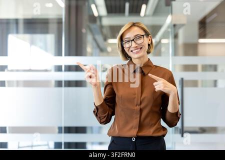 Donna d'affari sicura di sé con occhiali e camicia marrone, in piedi in un ufficio moderno, sorride e indica attivamente un'area vuota per la pubblicità Foto Stock