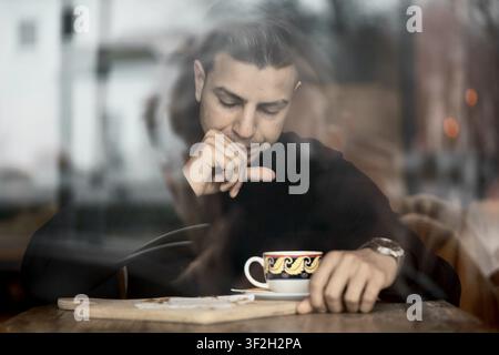 Soffocato da qualcosa, uomo in posa pensierosa a un tavolo da caffè con una tazza di caffè, visto attraverso una finestra riflettente, per far scendere una bevanda nella gola sbagliata Foto Stock