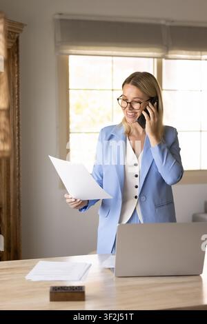 Donna d'affari che esamina documenti commerciali e parla al telefono in ufficio Foto Stock