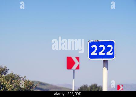 Segnale stradale blu che mostra il numero di strada 222. Un segnale direzionale rosso è visibile sullo sfondo. Cielo sereno sopra il paesaggio rurale Foto Stock