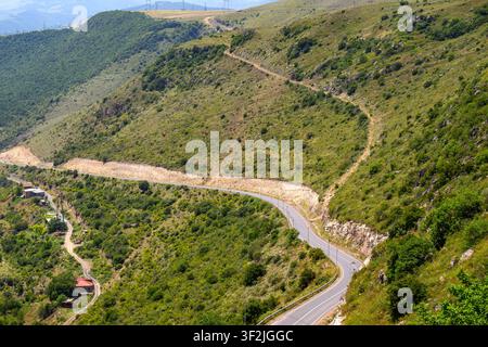 Strada tortuosa si snoda tra colline rigogliose e verdi. Un terreno roccioso delimita il percorso stretto asfaltato. Piccoletto villaggio incastonato nella valle sottostante Foto Stock