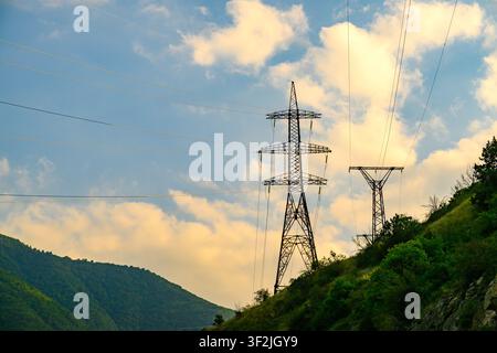 Alti tralicci elettrici si estendono su un paesaggio montuoso sotto un cielo parzialmente nuvoloso. Dolci colline verdi si innalzano dietro le torri Foto Stock