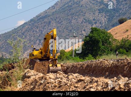 Escavatore giallo scava una trincea in terreno roccioso. Sfondo montuoso con alberi verdi Foto Stock