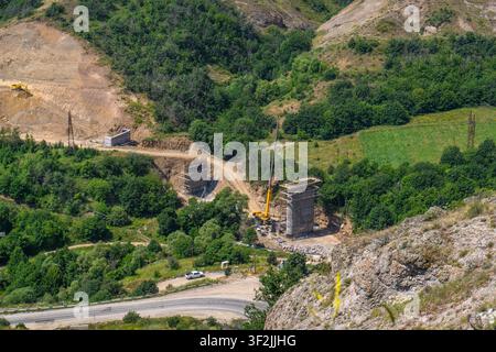 Grande gru per costruzioni solleva materiali vicino a vecchie rovine di pietra. Una foresta verde circonda la valle con una strada tortuosa. Sentieri sterrati collegano gli edifici Foto Stock