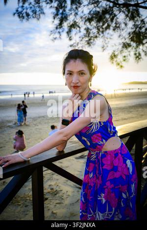 Una donna in abito floreale si appoggia a una ringhiera di legno sopra una spiaggia al tramonto a Putatan, Sabah, Malesia. Foto Stock