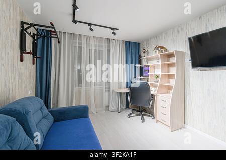 Camera accogliente con un divano blu, una scrivania con un computer, una libreria e una barra di trazione montata a muro. La luce naturale filtra attraverso la finestra Foto Stock