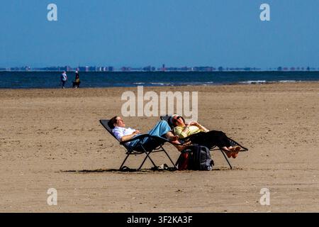 Due donne che si rilassano su sedie a sdraio pieghevoli sull'ampia distesa sabbiosa della spiaggia di Ainsdale facendo il bagno di sole con lo skyline lontano di Blackpool sullo sfondo. Southport, Regno Unito. Foto Stock