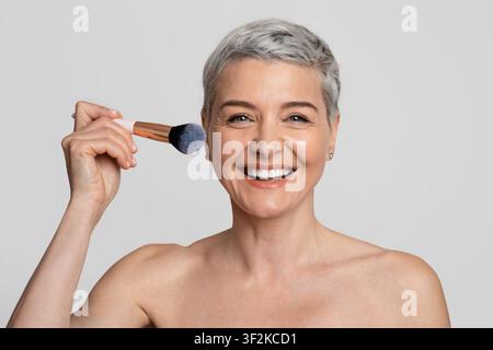 Bella donna sorridente di mezza età che fa il trucco, applicando il blush alla sua guancia Foto Stock