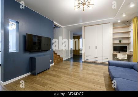 Soggiorno moderno con un muro accentato blu, TV montata a parete, pavimento in legno e un'anticipazione di una scala. Design minimalista con armadio bianco e giallo C Foto Stock