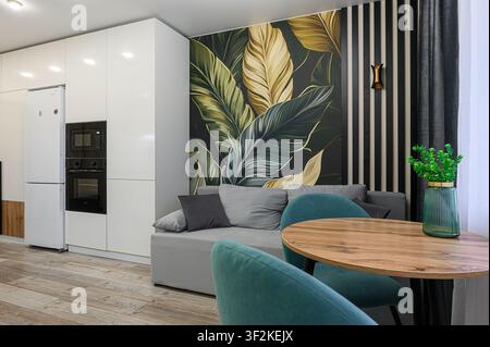 spazio abitativo moderno con un angolo cucina, caratterizzato da un divano grigio, un tavolo rotondo in legno, sedie turchesi e un murale botanico Foto Stock