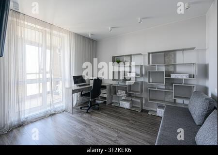 Stanza luminosa e moderna con una scrivania e un computer vicino a una finestra con tende trasparenti. Sono presenti una libreria grigia e un divano, creando uno spazio di lavoro minimalista Foto Stock