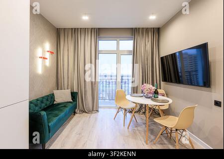 Soggiorno accogliente con un divano verde, zona pranzo vicino alla finestra e una TV elegante. Interno di appartamento moderno e stiloso Foto Stock