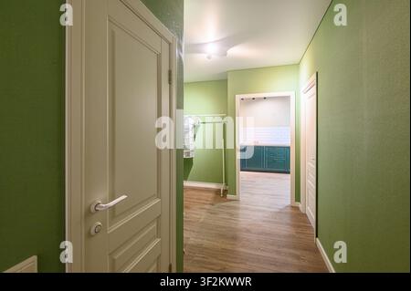 Corridoio dipinto in tonalità di verde con pavimenti in legno. A sinistra c'è una porta color crema. In lontananza si vede una cucina con mobili verde scuro Foto Stock