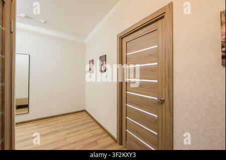 corridoio moderno con pavimento in legno, carta da parati beige chiaro e una porta in legno elegante con strisce bianche. Quadri sono appesi al muro. Uno specchio è visibile Foto Stock