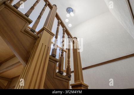 Scatto a basso angolo mostra una scala in legno con ringhiere ornate. Sopra, un moderno lampadario pende da un soffitto bianco strutturato, completando il ro… Foto Stock