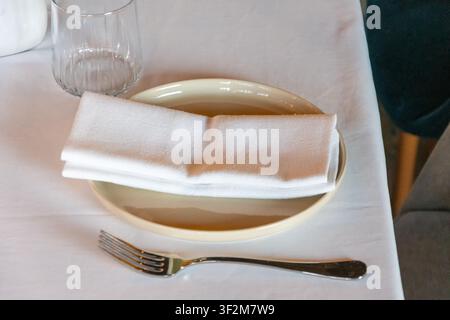 Impostazione minimalista del tavolo da ristorante con tovagliolo di stoffa bianco, forchetta, bicchiere da bere e piatto di ceramica in primo piano. Foto Stock