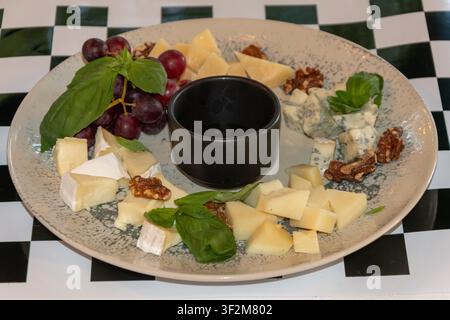 Vassoio di formaggi assortiti con uva, noci, basilico e salsa da intingere servito per la cena in un ristorante gourmet. Foto Stock