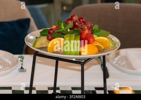 Vassoio di frutta assortita fresca con uva, mele, arance, fragole e menta servito in un elegante interno di ristorante. Foto Stock