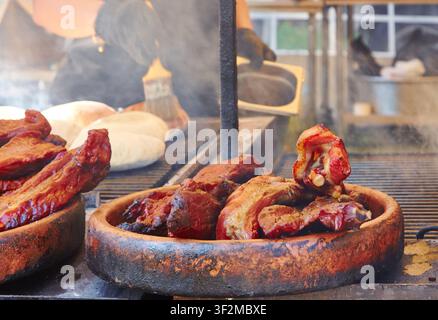 Costine e bistecche grigliate su un barbecue con fumo che si alza, mentre uno chef prepara ulteriori piatti sullo sfondo di un impianto di cottura all'aperto Foto Stock