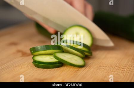 Tagliare i cetrioli con un coltello grande su un tagliere di legno a fuoco ravvicinato. Una donna prepara un'insalata di verdure fresca a casa Foto Stock