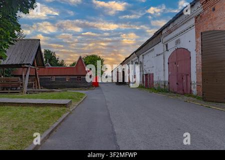 Cortile di fattoria storica con fienili tradizionali, edifici in legno rustici e vecchi magazzini agricoli lungo una strada lastricata tranquilla. Muri di mattoni bianchi, scoloriti Foto Stock