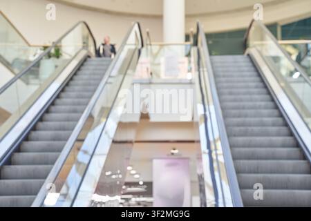 Coppia di scale mobili che fiancheggiano la scala centrale vuota all'interno dell'atrio luminoso di un centro commerciale con ringhiere in vetro e acquirenti visibili al livello superiore in messa a fuoco morbida. Foto Stock