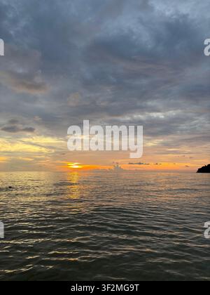 Trascinante tramonto arancione che si apre tra le nuvole di tempesta sulle acque calme del mare in Cambogia, con riflessi dorati sulla superficie increspata dell'acqua. Aprile 2025. Foto Stock