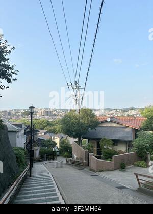 Kanazawa, Giappone – Quartiere residenziale tradizionale con case in legno, vegetazione rigogliosa, linee elettriche e vista panoramica sulla città in una limpida mattina estiva. Foto Stock