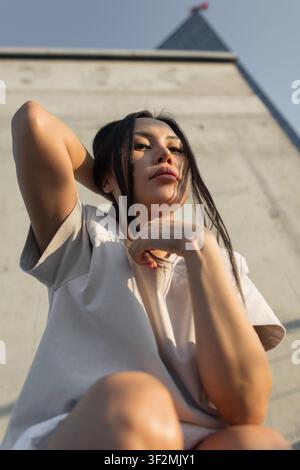 Ritratto in basso angolo di una giovane donna asiatica sicura di sé che guarda la telecamera contro un muro di cemento nella luce calda del pomeriggio, tenendo i suoi lunghi capelli scuri Foto Stock