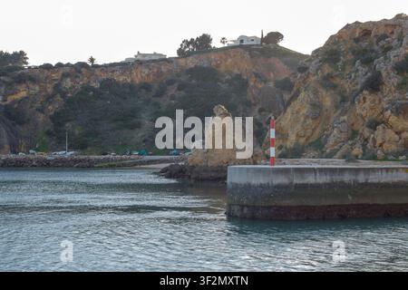 La scena mostra una costa rocciosa al tramonto con un molo di cemento. Un faro si erge alla fine del molo. Case sono visibili sulla collina nella ba Foto Stock