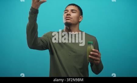 Giovane che assaggia succo verde da una piccola bottiglia, mano a bocca mentre tiene la bottiglia sullo sfondo dello studio di tè; calma benessere. Foto Stock